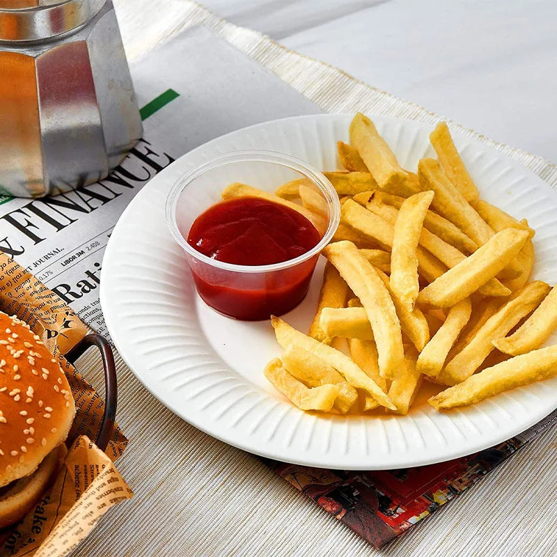 A plate of french fries and a small cup of red sauce on a white plate with newspaper in the background.