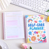 A colorful self-care planner notebook with stickers and a white journal lying next to a laptop keyboard.