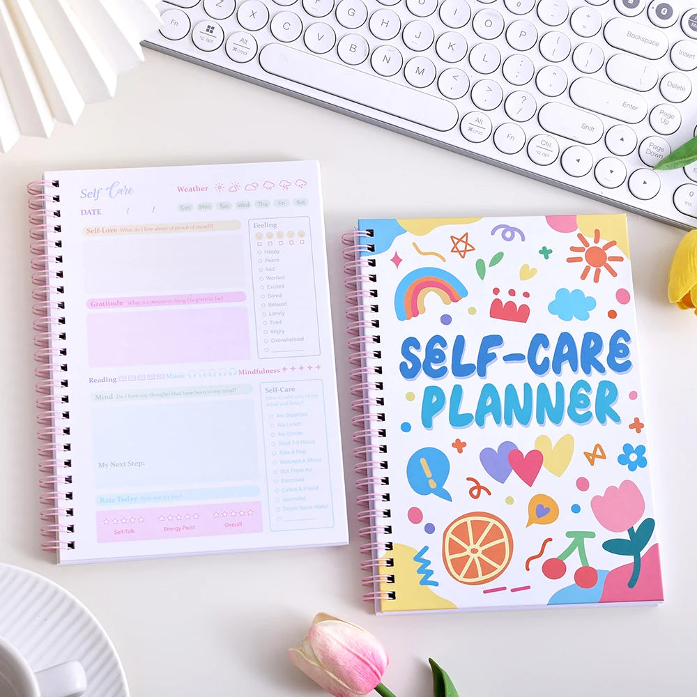A colorful self-care planner notebook with stickers and a white journal lying next to a laptop keyboard.