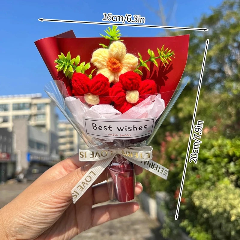 A hand holds a bouquet of handmade crochet flowers featuring red and yellow blooms wrapped in clear cellophane.
