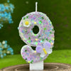 A white number cake candle shaped like the number 8 decorated with purple and yellow flowers and a purple butterfly