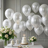 A display of pearl white latex balloons, roses, and a tiered cake on a white table for a wedding or party.