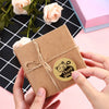 Hands are holding a brown kraft paper gift box tied with twine and a thank you sticker on a pink background.