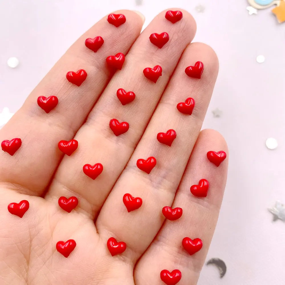 A hand displays numerous small red heart-shaped rhinestones for nail art and crafts.