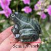 Une main tient une figurine miniature de grenouille de jade vert foncé contre un arrière-plan vert flou.