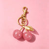 Gold chain charm featuring two pink glitter glass stones shaped like cherries on a light pink background.
