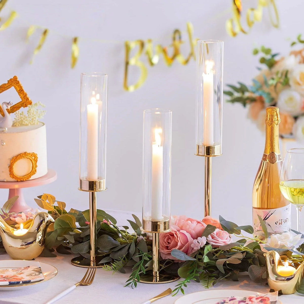 A display of three tall glass hurricane candle holders with gold accents and pink roses on a table.