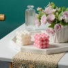 A set of two white and pink cube soy wax candles displayed on a white tray with a glass bottle in a teal background.