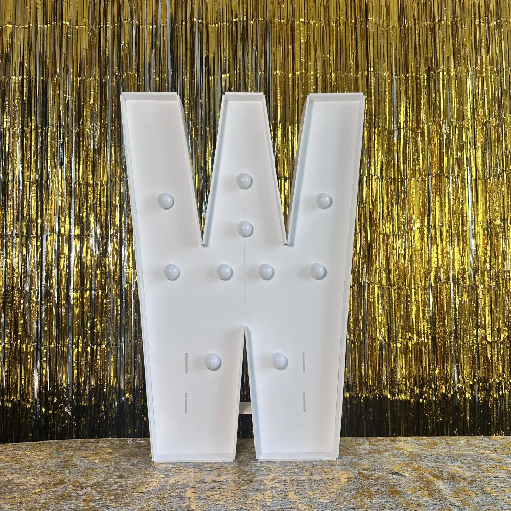 A large white marquee light-up letter W stands against a shimmering gold curtain backdrop.