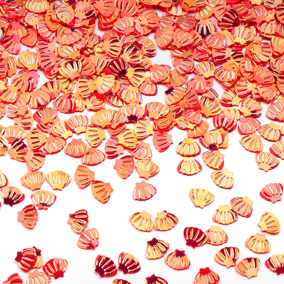 A close-up of small, orange and pink sea shell PVC sequins scattered on a white background for nail art and craft decoration.