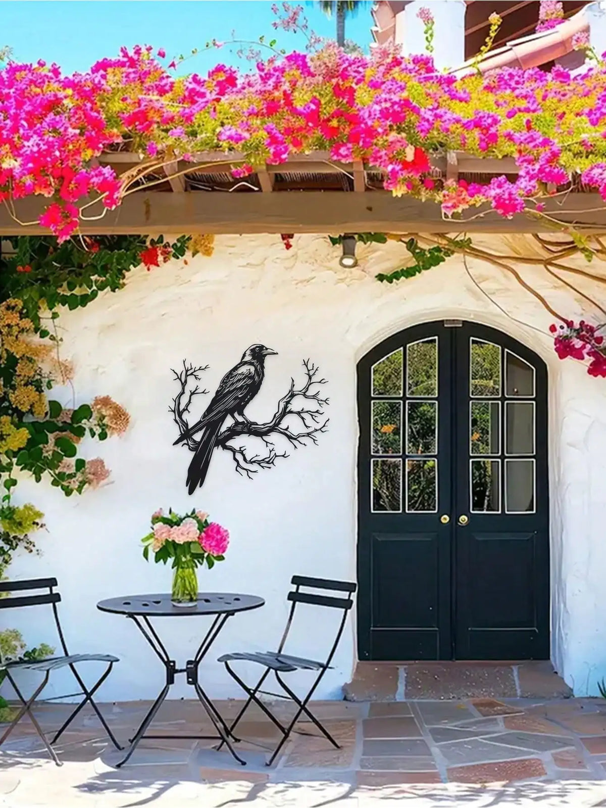 Black raven metal wall art mounted on a white stucco wall under pink flowering vines.