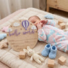 A baby lying on a blanket next to a wooden birth announcement plaque with the words hello world and various small wooden toys