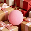 A pink velvet flocked Christmas ornament with a gold hanger resting on a stack of red and white wrapped gifts.