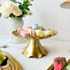 A golden plastic cake stand holds colorful macarons and white roses on a white surface.