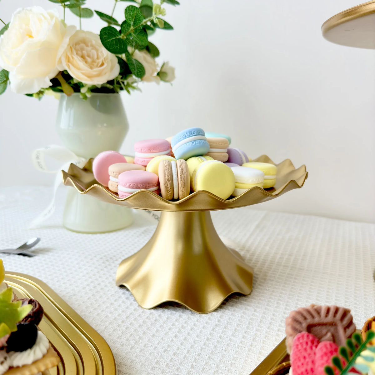 A golden plastic cake stand holds colorful macarons and white roses on a white surface.