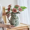 A hand holds a rustic ceramic milk pitcher with a cracked glaze and pastoral design, filled with pink roses and green foliage