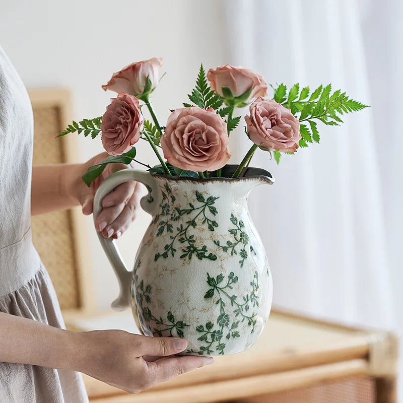 A hand holds a rustic ceramic milk pitcher with a cracked glaze and pastoral design, filled with pink roses and green foliage