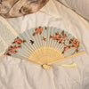 A light blue folding hand fan decorated with pink and brown floral designs rests on a white fabric.