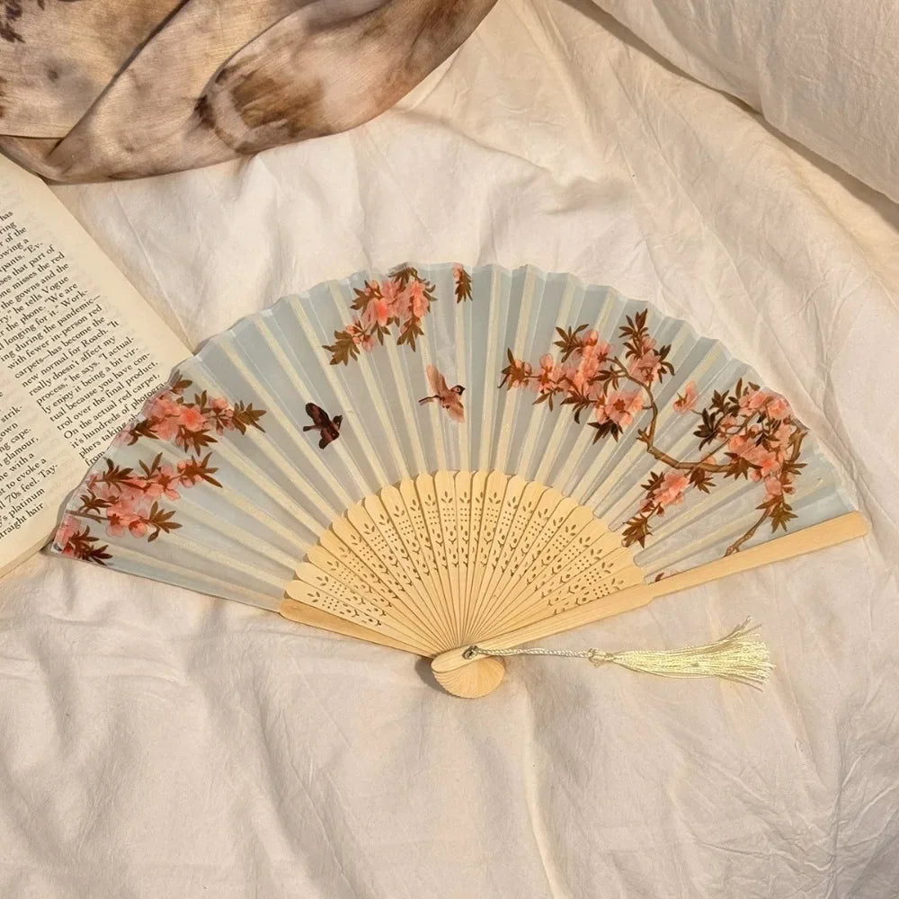 A light blue folding hand fan decorated with pink and brown floral designs rests on a white fabric.
