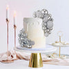 A white tiered cake decorated with silver disco ball toppers and lit candles on a gold stand.