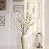 A vase holds a branch of white faux plum blossoms with stems, set in a bright indoor setting.