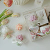 A clear glass box containing a pink tulip candle and other floral elements on a white surface.