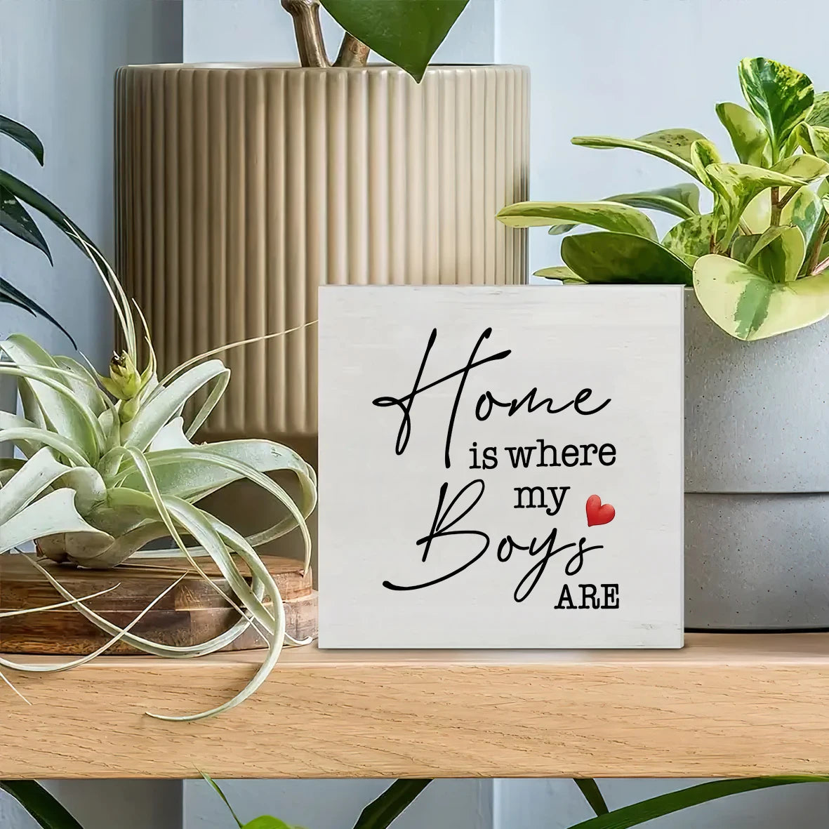 A rustic wooden sign with black script that reads Home is where my boys are with a small red heart, displayed on a light wood