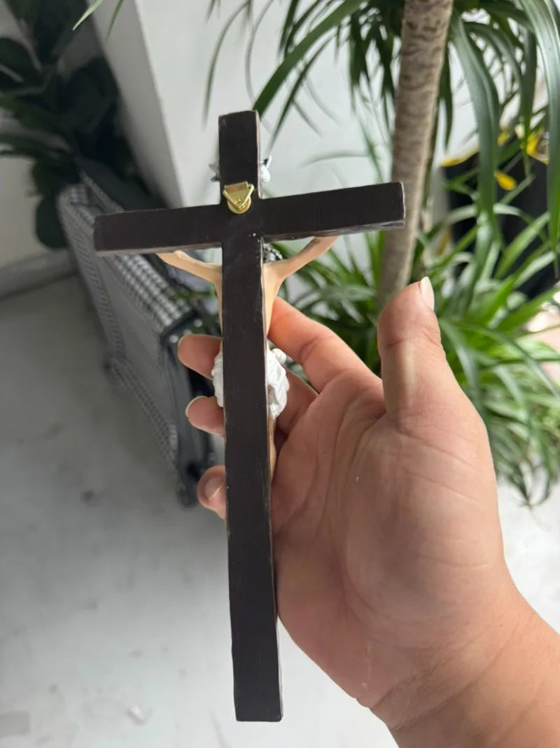 A hand holds a dark wood crucifix with a gold-colored finial against a blurred green plant background.