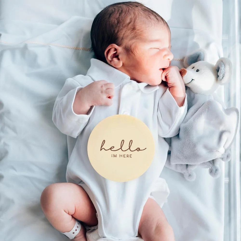 A newborn baby lying on white fabric, holding a wooden sign that reads Hello I'm Here.