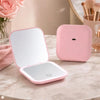 A pink portable folding LED makeup mirror with a reflective surface and a separate magnifying base is displayed on a marble t