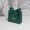 A dark green paperboard thank you gift bag tied with a satin ribbon, resting on a white surface.