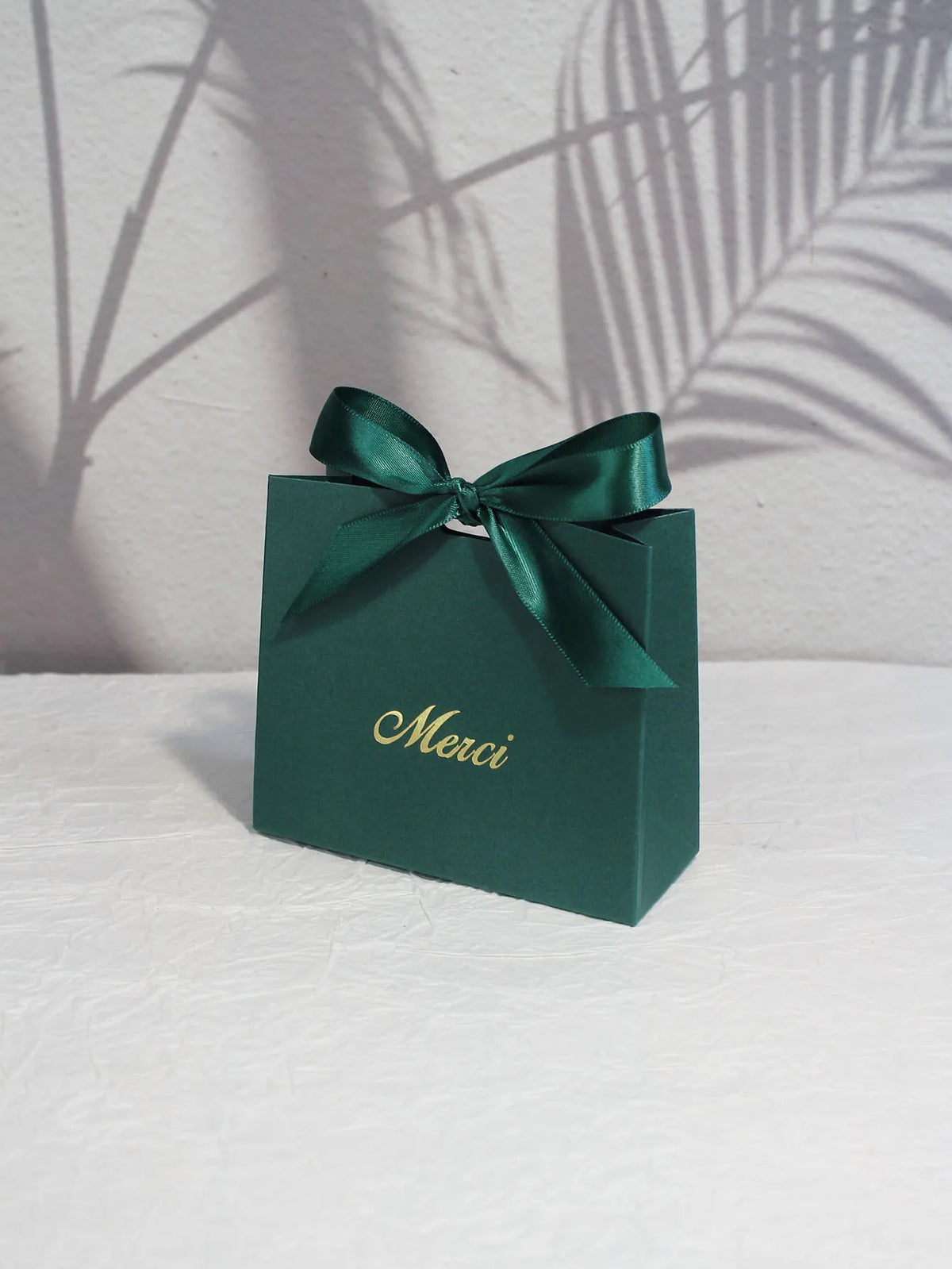 A dark green paperboard thank you gift bag tied with a satin ribbon, resting on a white surface.