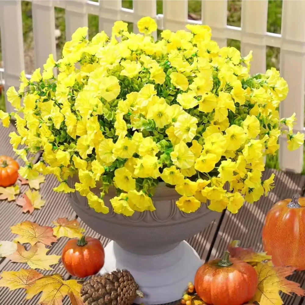 A vibrant bouquet of artificial yellow flowers displayed in a gray urn surrounded by autumn leaves and pumpkins.