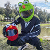 A green cartoon plush helmet cover with large eyes is shown outdoors.