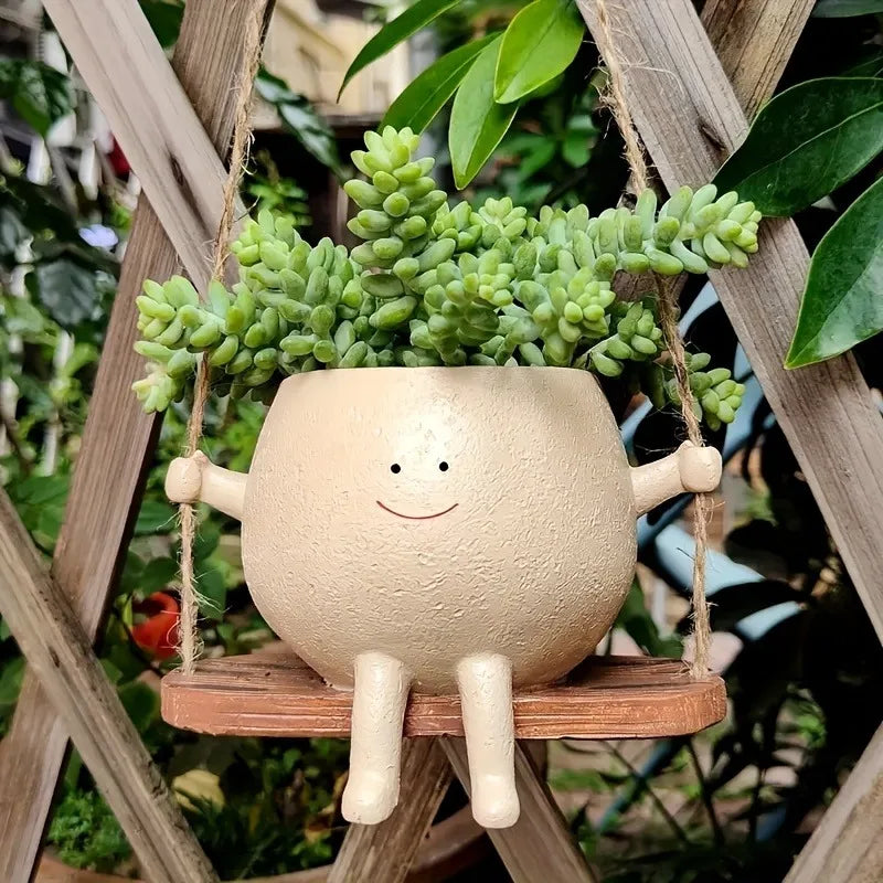 A light beige resin succulent planter with a round face is hanging from a wooden swing base amidst green foliage.