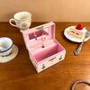 A pink ballet music box with a hinged lid displaying rotating figurines and jewelry storage, next to a teacup and a strawberr