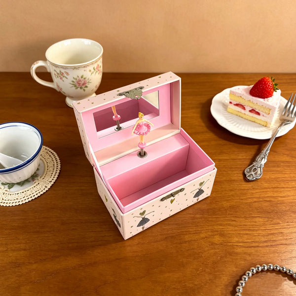 A pink ballet music box with a hinged lid displaying rotating figurines and jewelry storage, next to a teacup and a strawberr