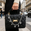 A black quilted leather handbag with a gold chain and charms is held by a person in a black coat on a city street.