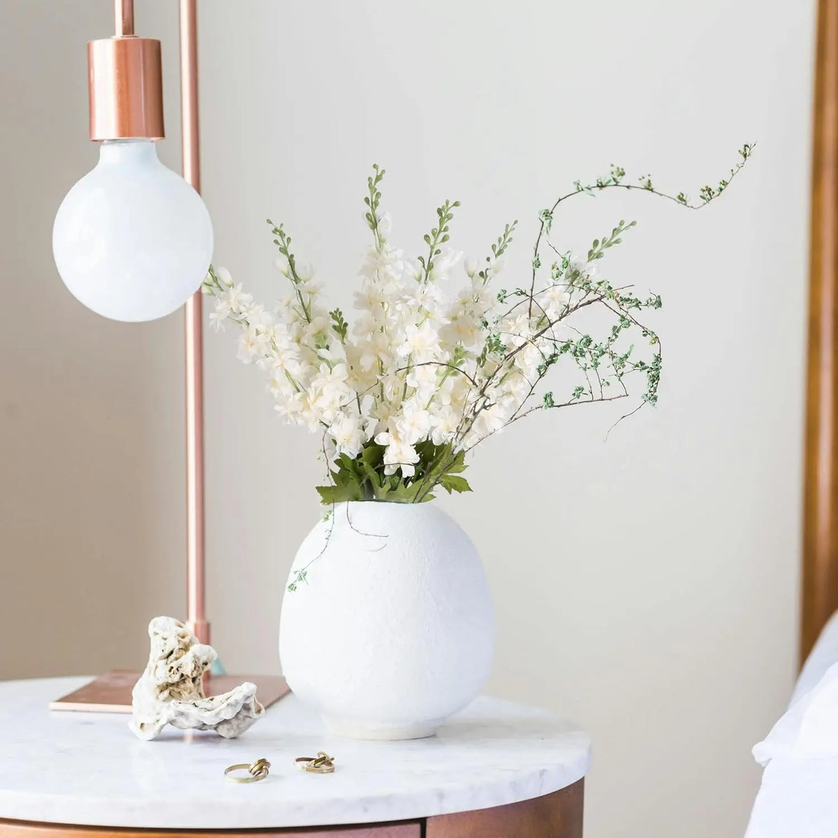Un vase de fleurs artificielles de delphinium blanches sur une table en marbre à côté d'une lampe dorée.