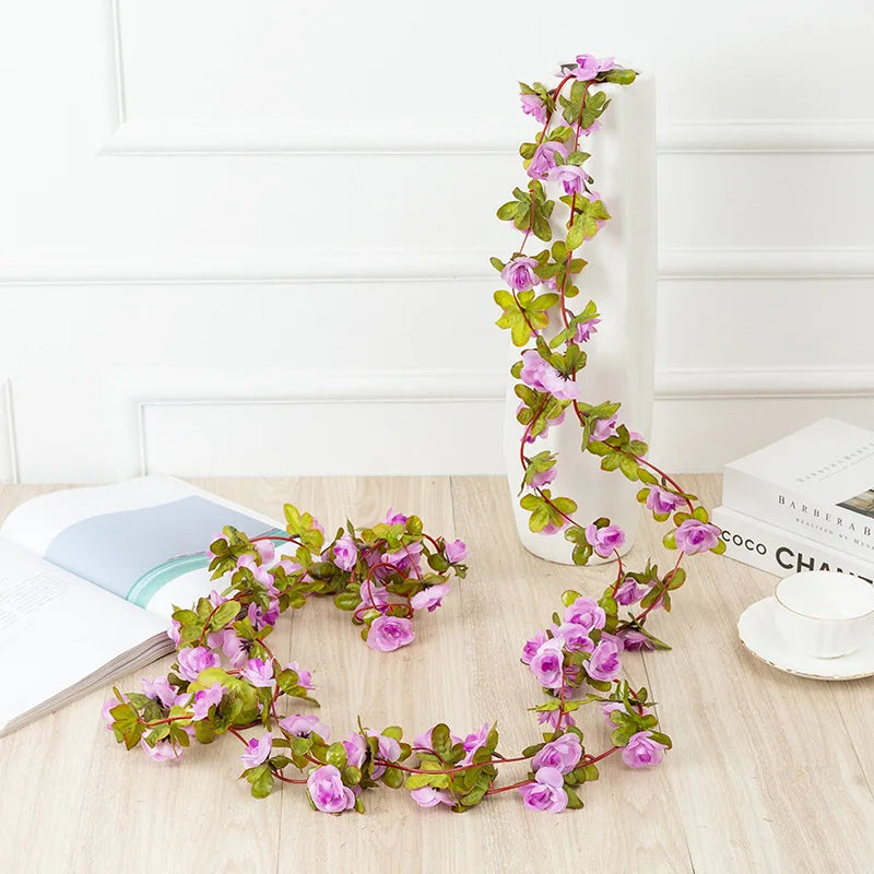 A silk rose garland with pink flowers is displayed against a white wall and next to a white rectangular object.