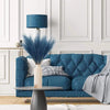 A teal lamp with a blue shade sits on a sofa, decorated with artificial pampas grass stems.