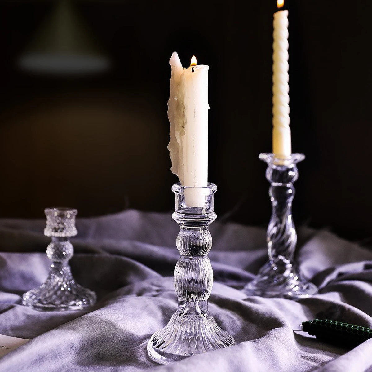 A tall white pillar candle in an elegant clear glass holder sits on a gray fabric surface.