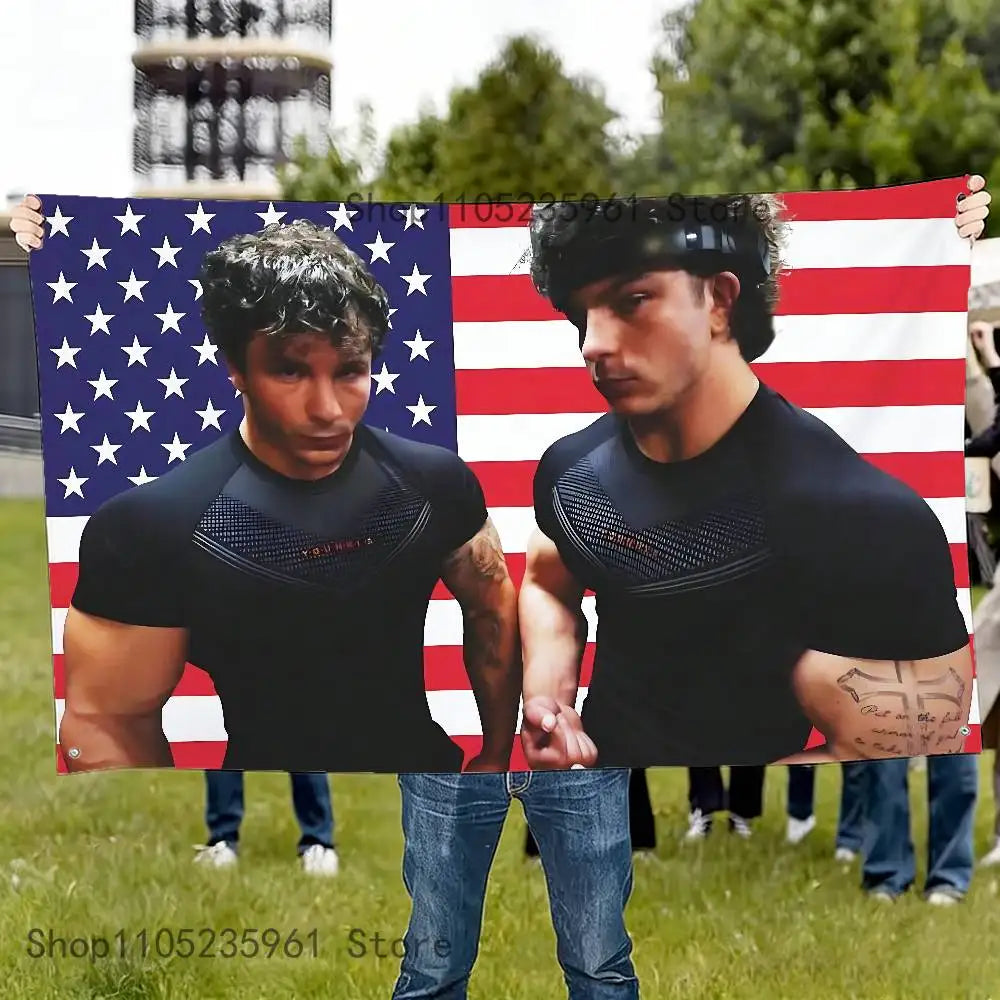 A muscular man wearing a black shirt stands in front of an American flag banner with red and white stripes.