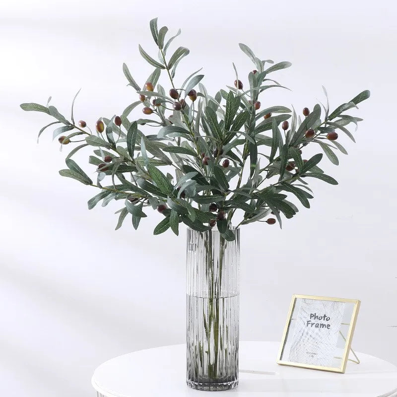An artificial olive branch with dark green leaves and small reddish-brown fruits displayed in a clear glass vase.