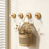A woven straw basket with a rope handle is hanging on a light-colored wall rack with four wooden hooks.