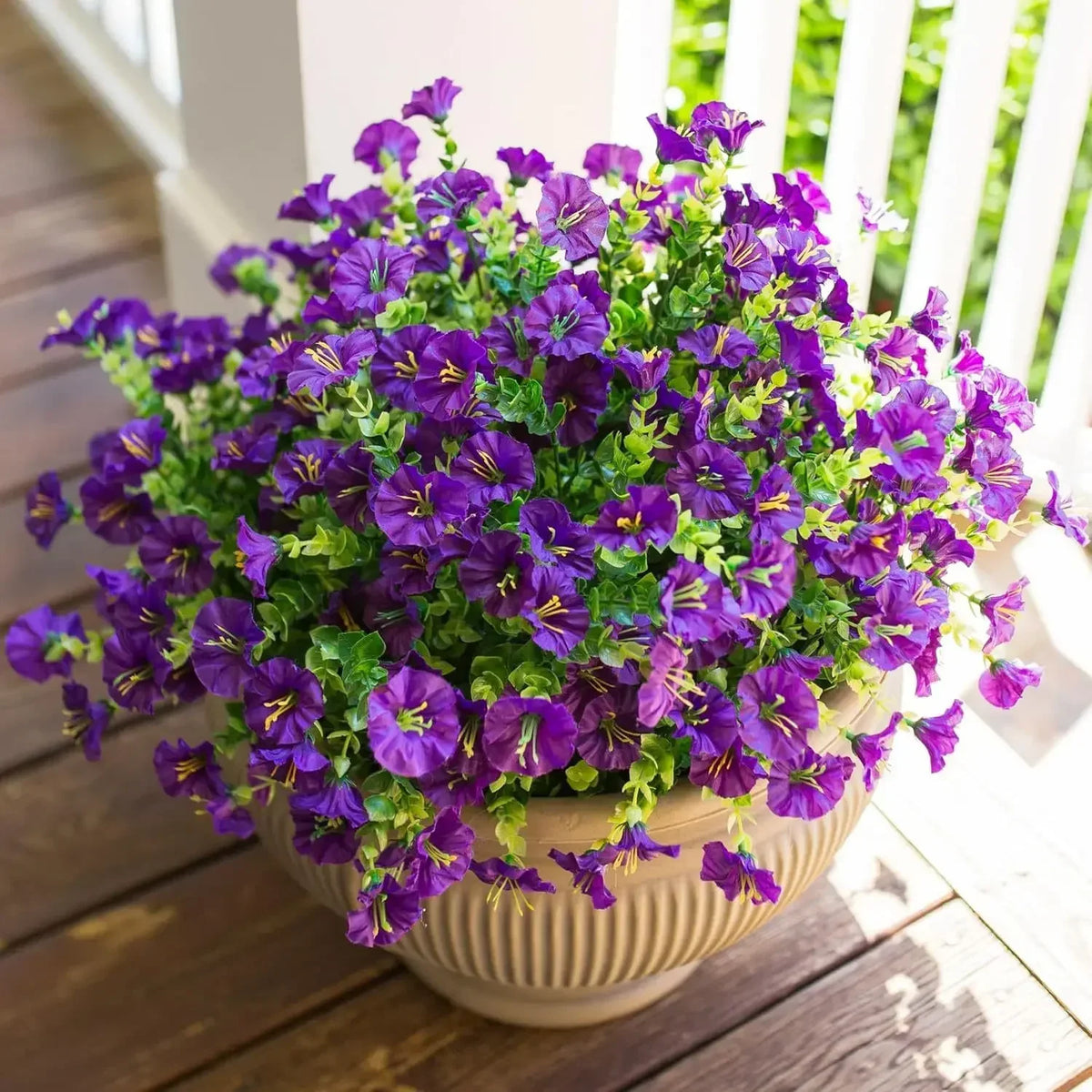 A potted arrangement of purple artificial morning glory flowers and green eucalyptus stems on a wooden deck.