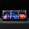 Three rough crystal specimens in clear acrylic boxes display purple, blue, and red colors.