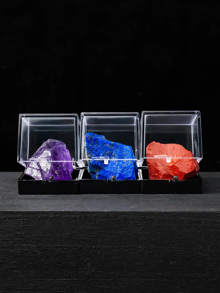 Three rough crystal specimens in clear acrylic boxes display purple, blue, and red colors.