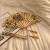 A beige and green leopard print folding hand fan with a black handle resting on a light-colored sheet.