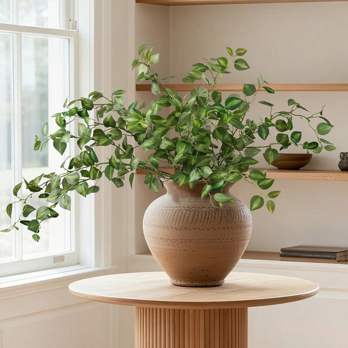A potted artificial eucalyptus stem with silk green leaves sits on a light wood table near a window.